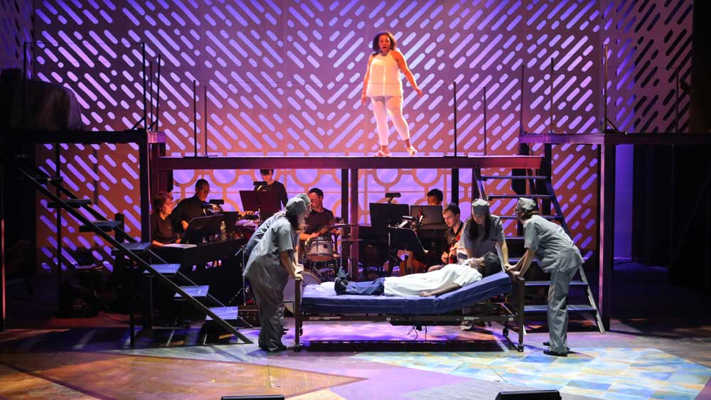 Female actress performing on an elevated stage with an actor on a hospital bed below surrounded by nurses