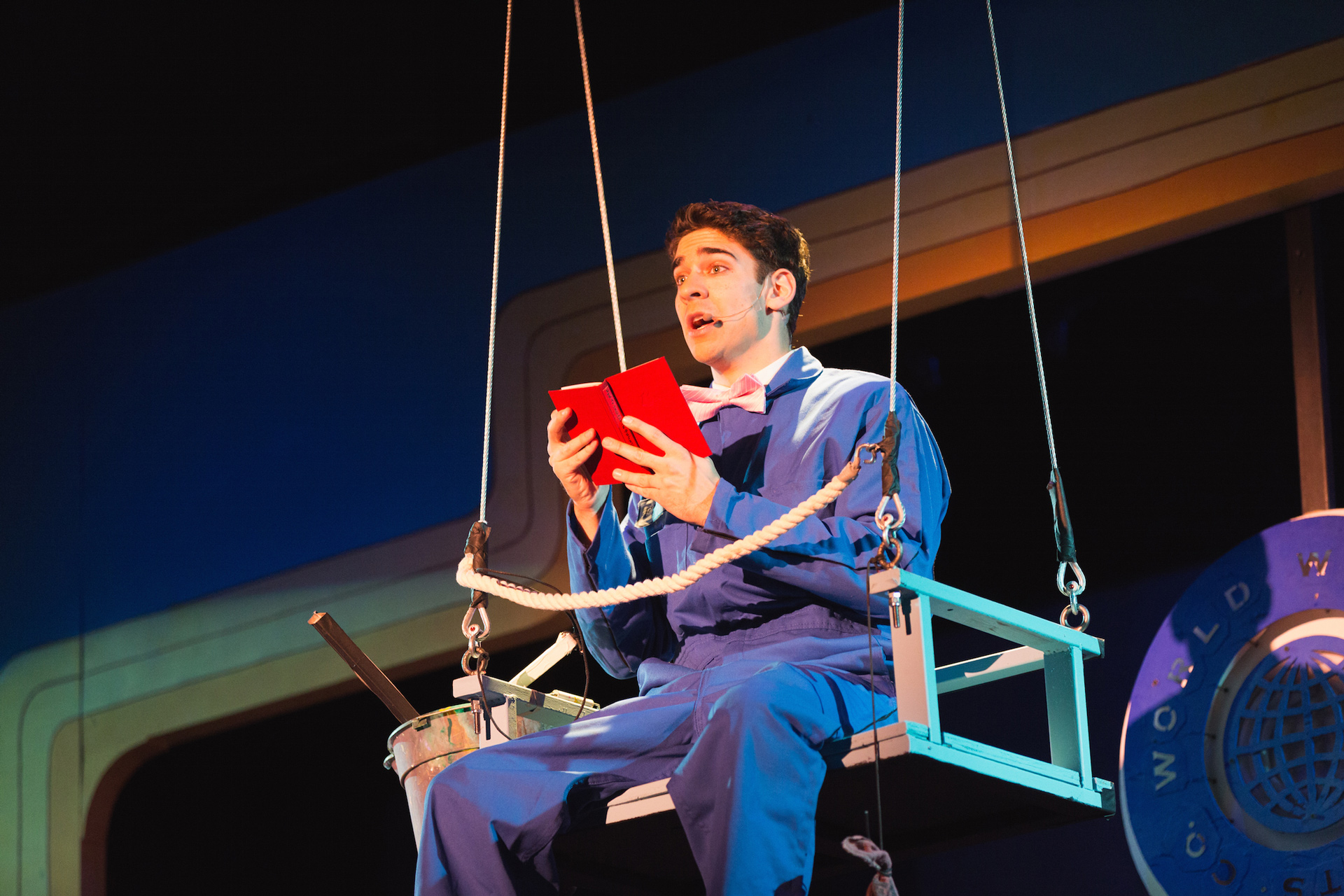 Man holding a red book and sitting in a chair connected to the ceiling by cables