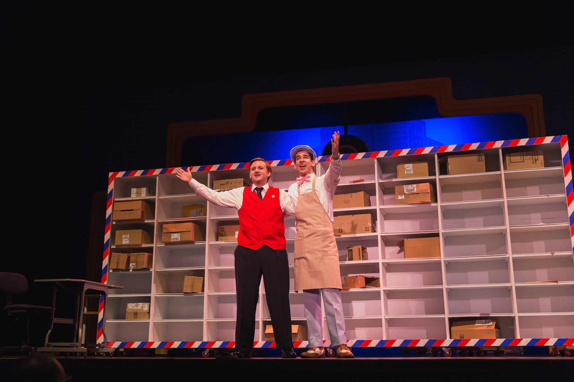 Two male actors holding their hands up with a wall of packages behind them