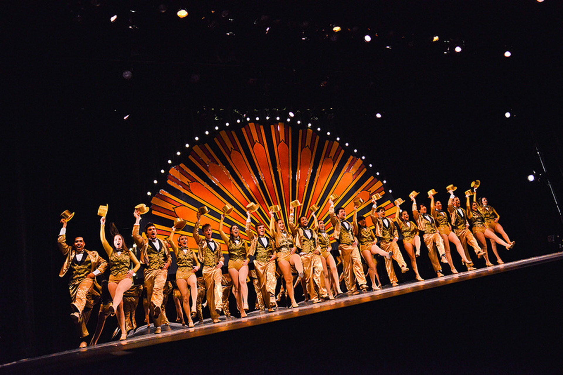 Actors and actresses line dancing with gold outfits and a starburst backdrop