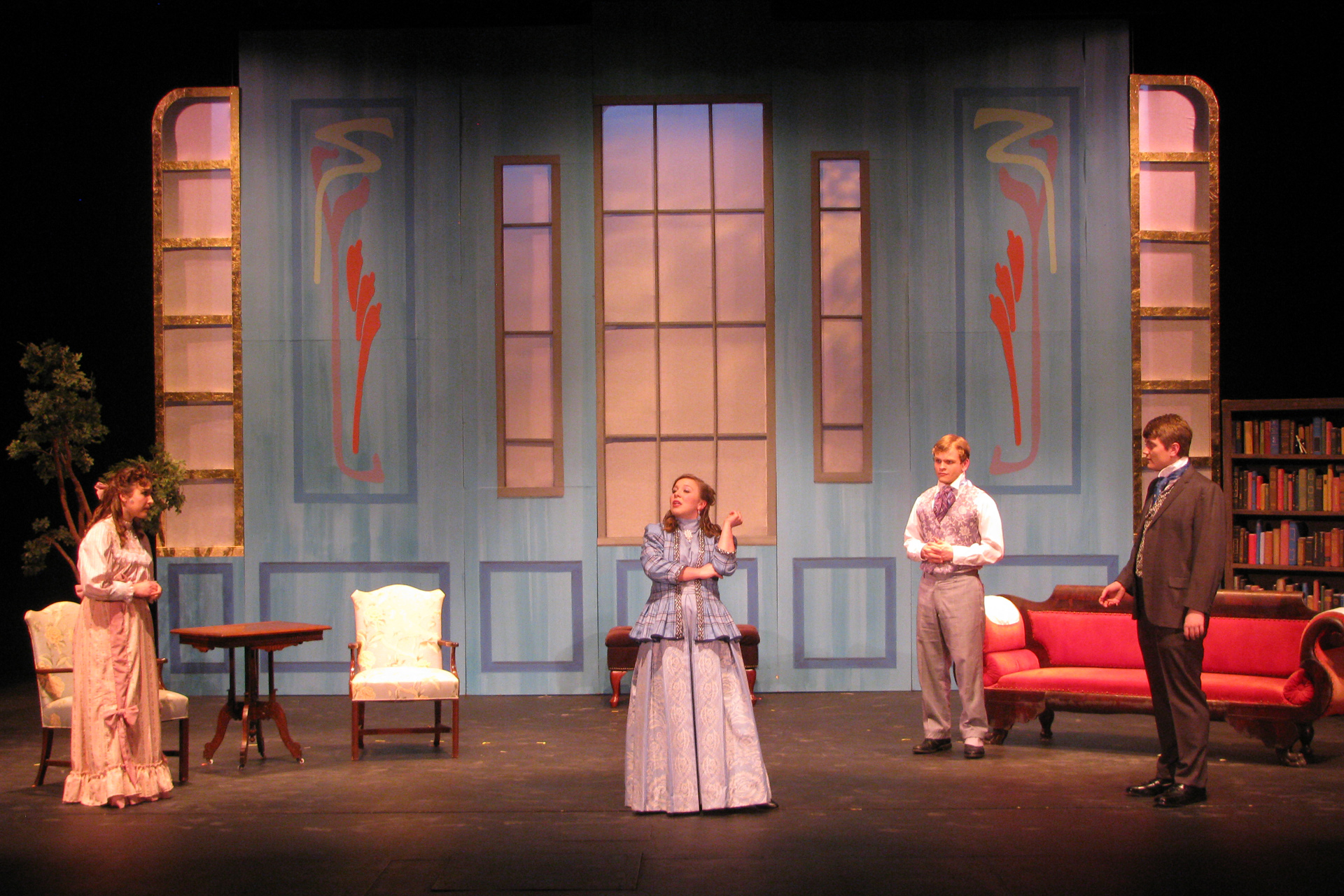 Actress wearing period attire stands in the middle of a victorian room with other actors