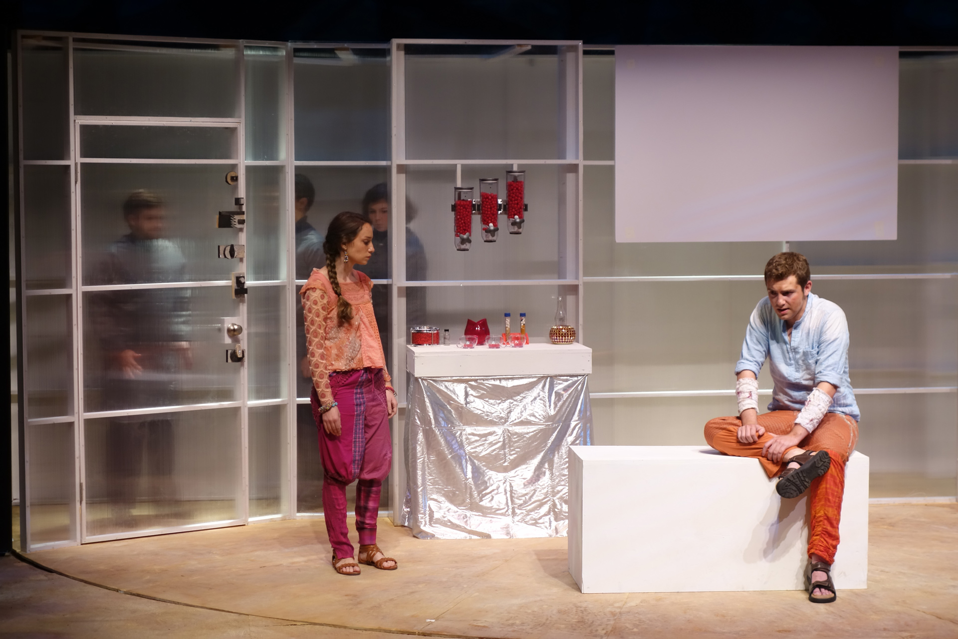 An actor sitting with a female actor standing near a semi-transparent doorway