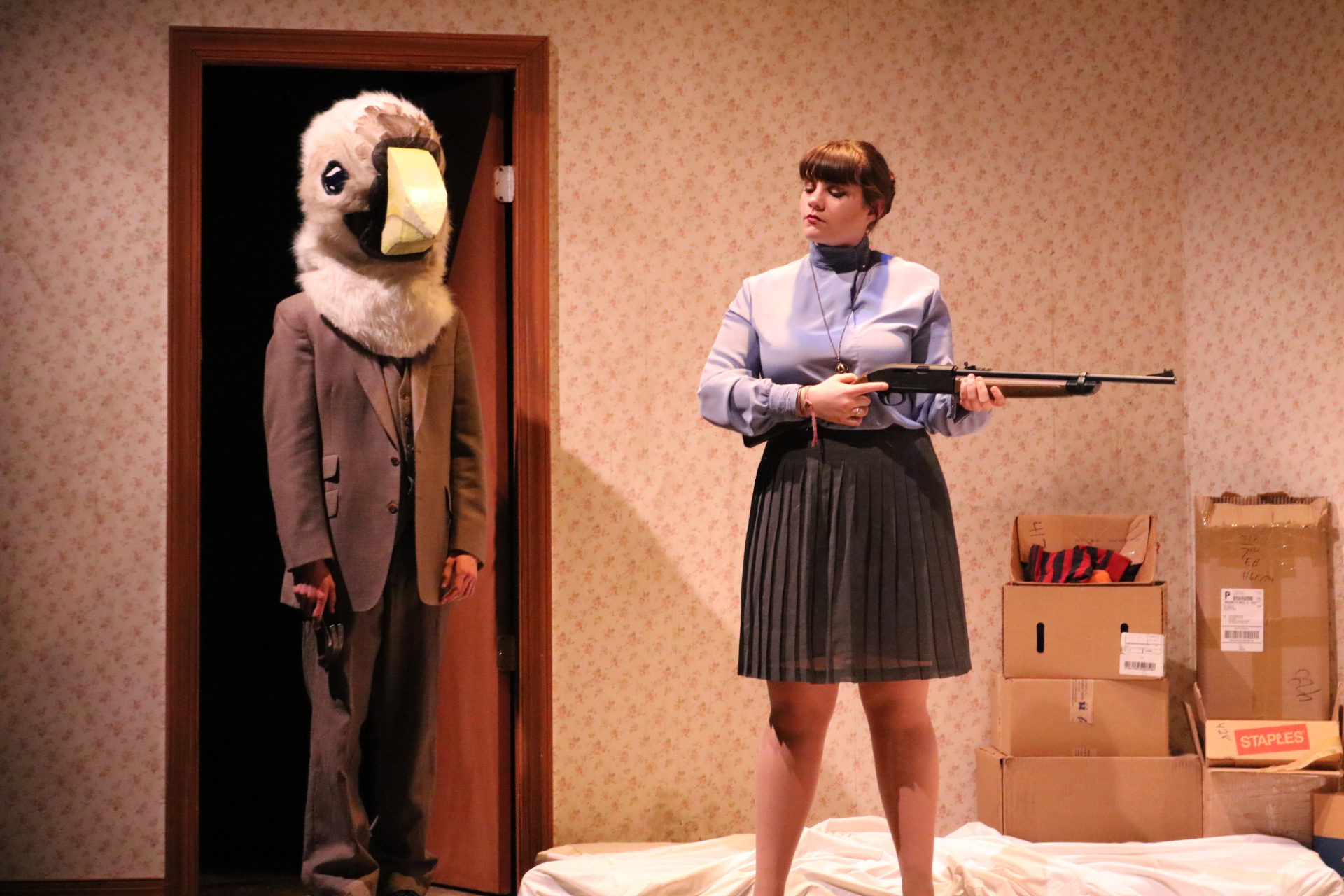 An actress holding a rifle with a man wearing a bird head costume standing near a doorway