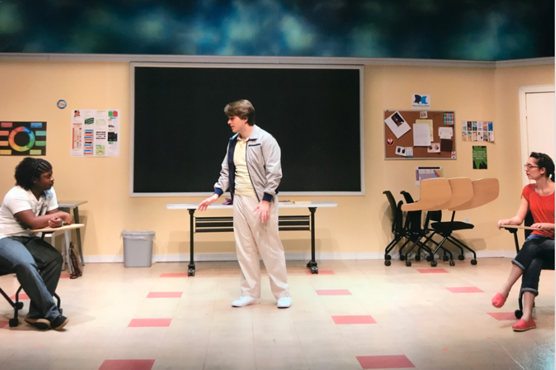 Male actor stands in front of a chalkboard speaking to students