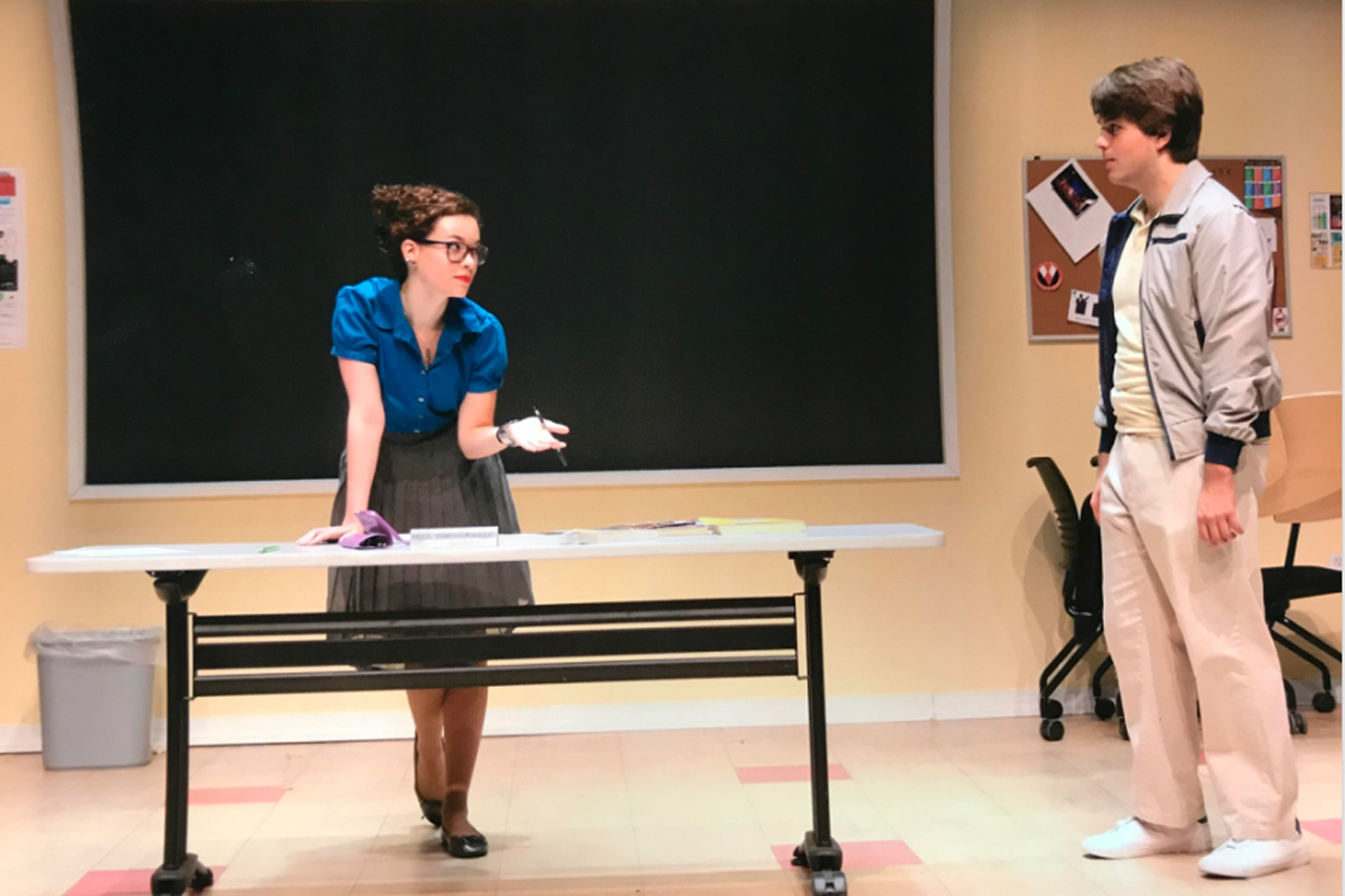 Actress dressed as a school teacher leaning on desk and speaking to another actor