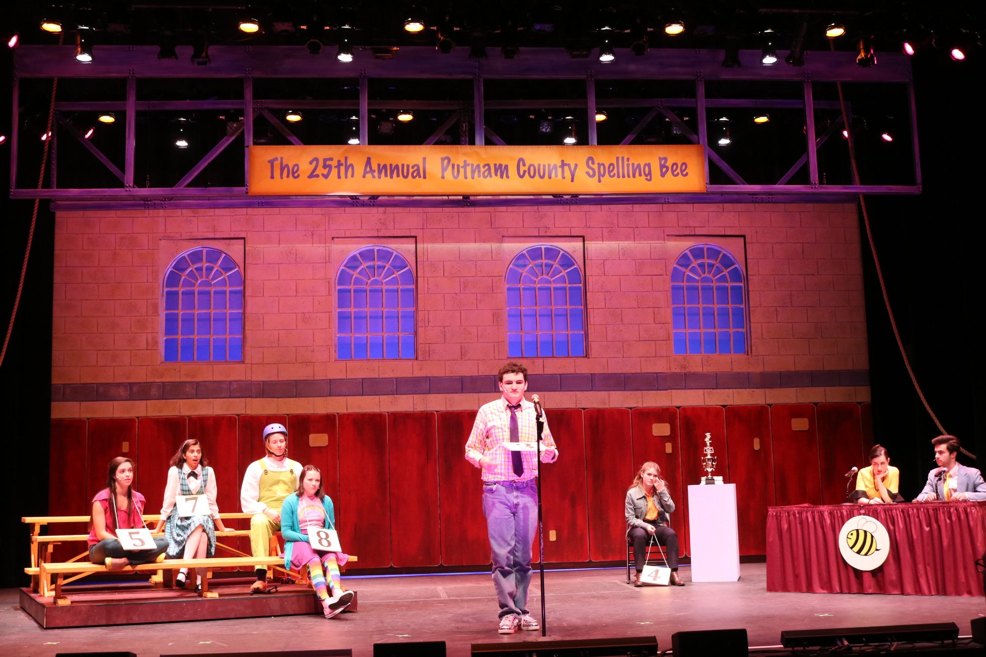 Man standing at a microphone while students sit on bleachers to the left at a spelling bee