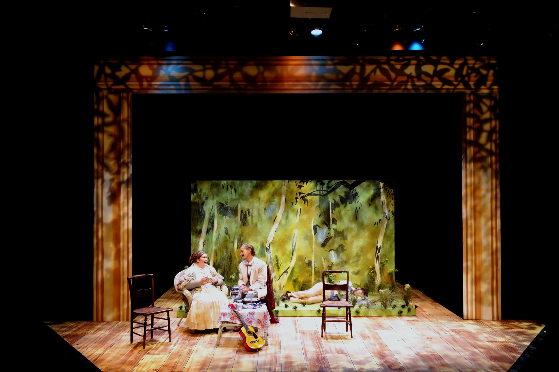 Older man and woman sitting at a table in period attire with a man laying on the ground behind them