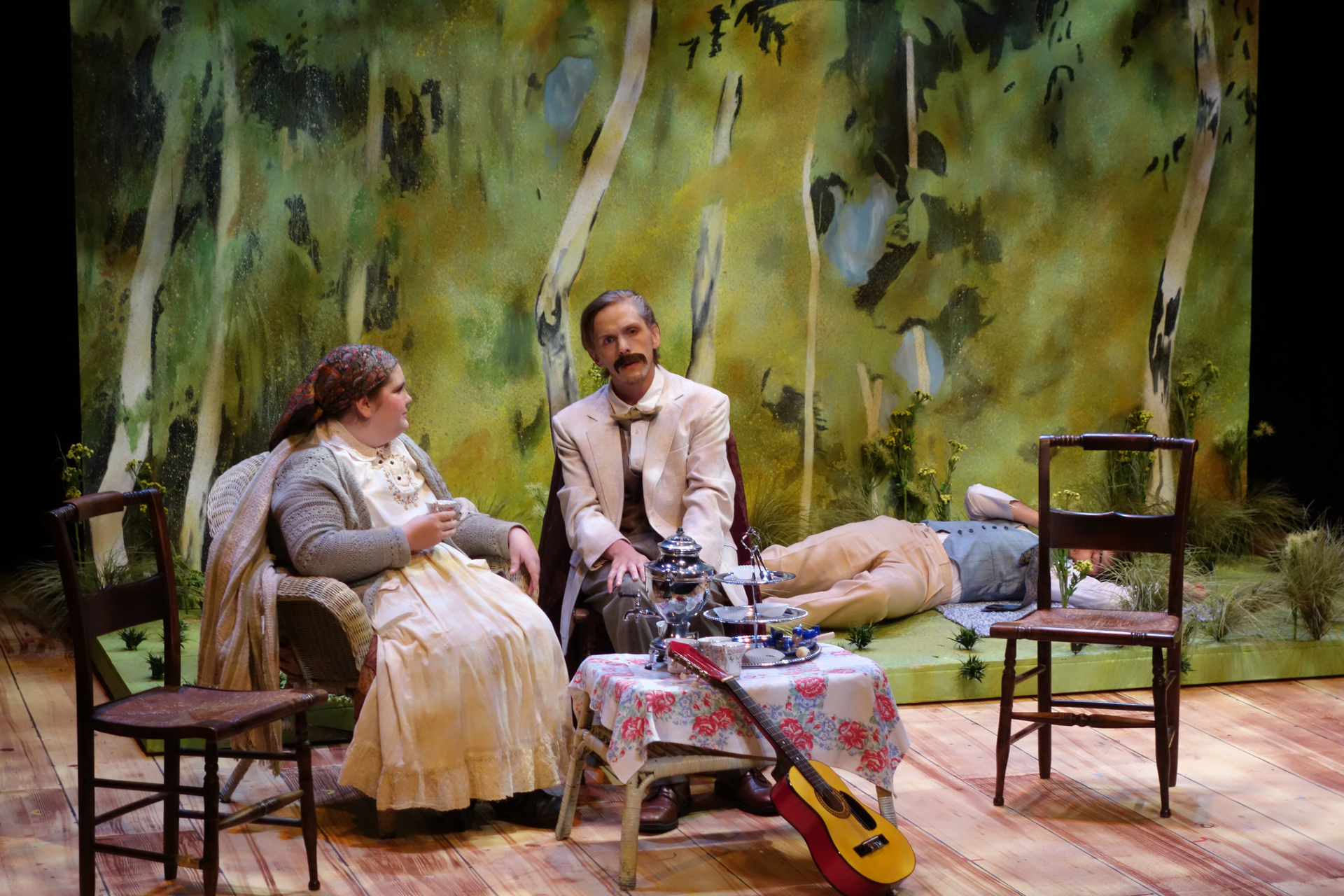 Close up of actor and actress sitting at a table with a man laying on the ground behind them