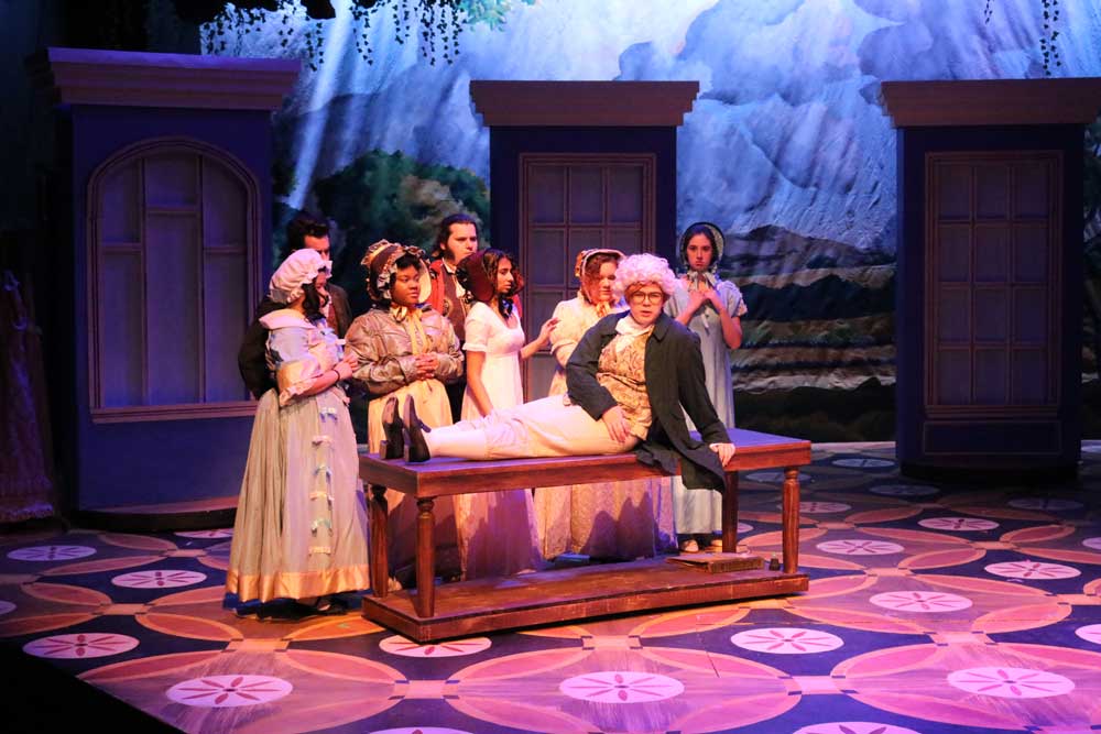 Male actor laying on a wooden table surrounded by female actresses