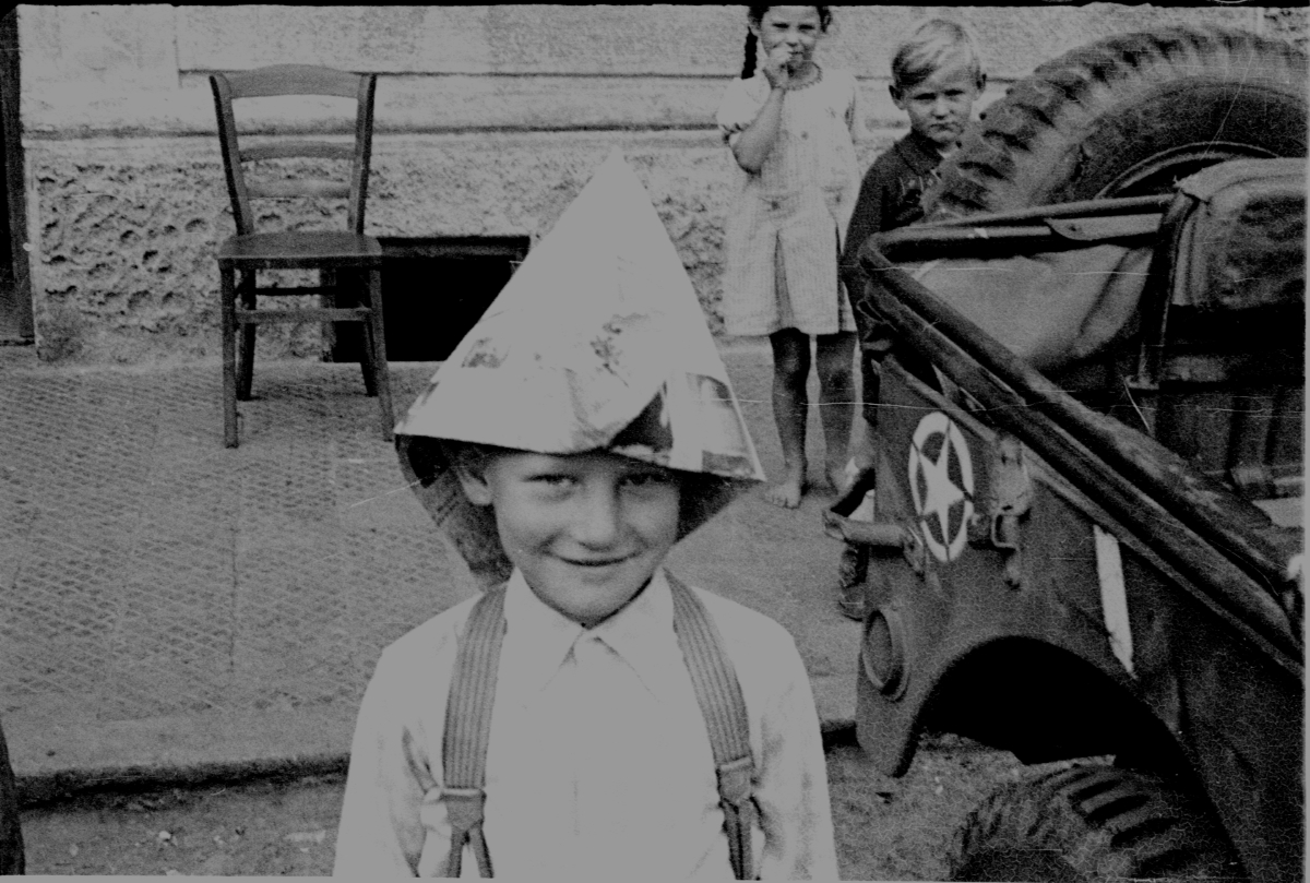 Little boy wearing newspaper hat standing next to an army jeep.