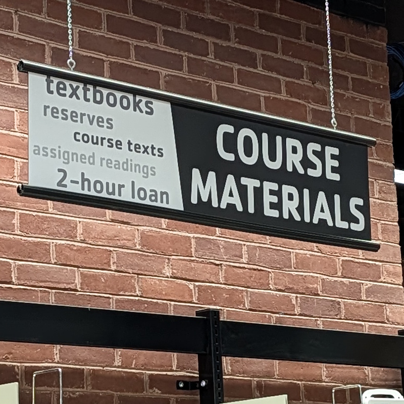 A black and white sign with Course Materials printed on it, hanging in front of a red brick wall.