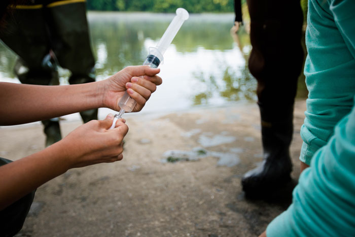 Person holding syringe filled with river water alongside a river