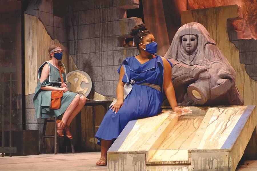 A woman sitting on a stage prop wearing a mask in the play Antigone Now