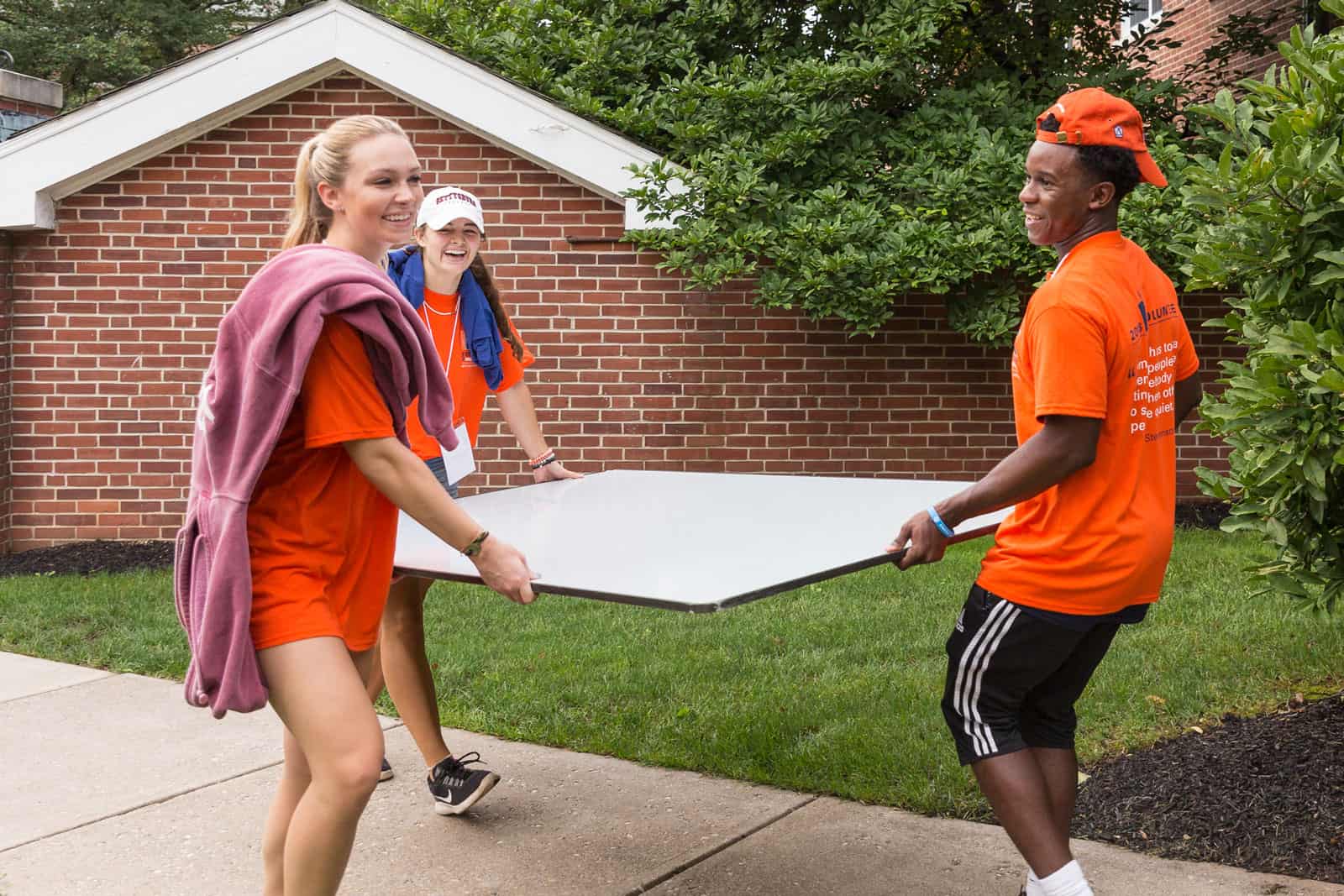 three smiling students in orange Move-In Day t-shirts carrying furniture
