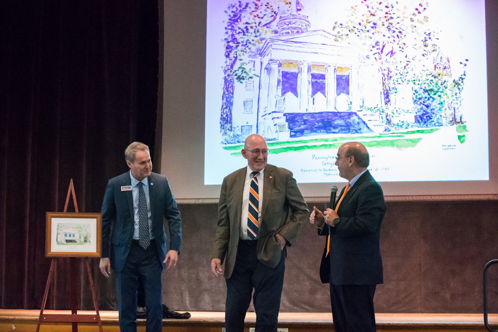President Bob Iuliano and two others stand before the audience, in front of a projected artist's rendering of Pennsylvania Hall