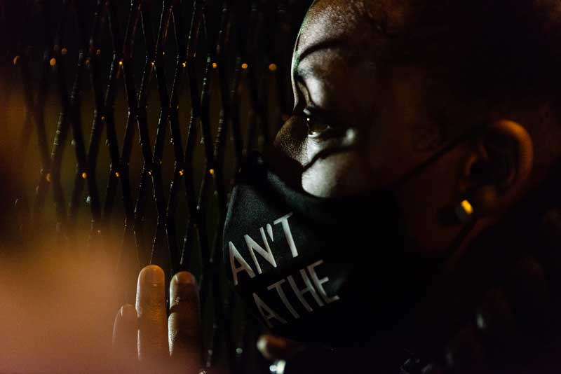 Demonstrator wearing a dark mask looking through a chain link fence