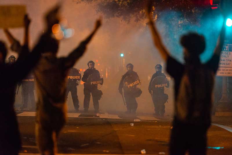 Secret Service officers in riot gear standing in a line formation in front of protestors raising their arms in the air