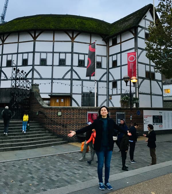 Julia stands with arms spread in front of the Globe Theatre