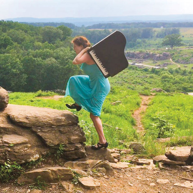 Jocelyn Swigger carrying a small piano on her back on the Gettysburg Battlefield