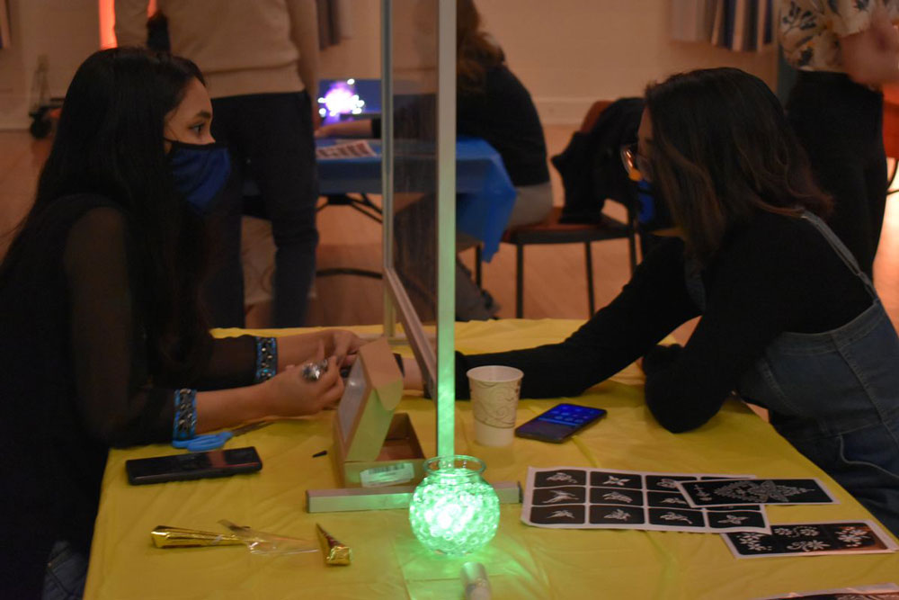 Pratikshya Prasai ’23 applying a henna design to another student's hand with a plexiglass barrier between them