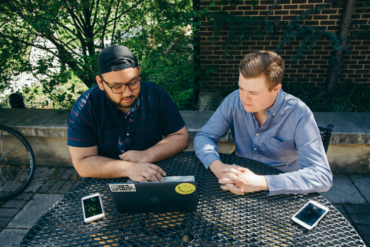 KKaran Shrestha and Jason Maltby sit outside while working on a laptop.