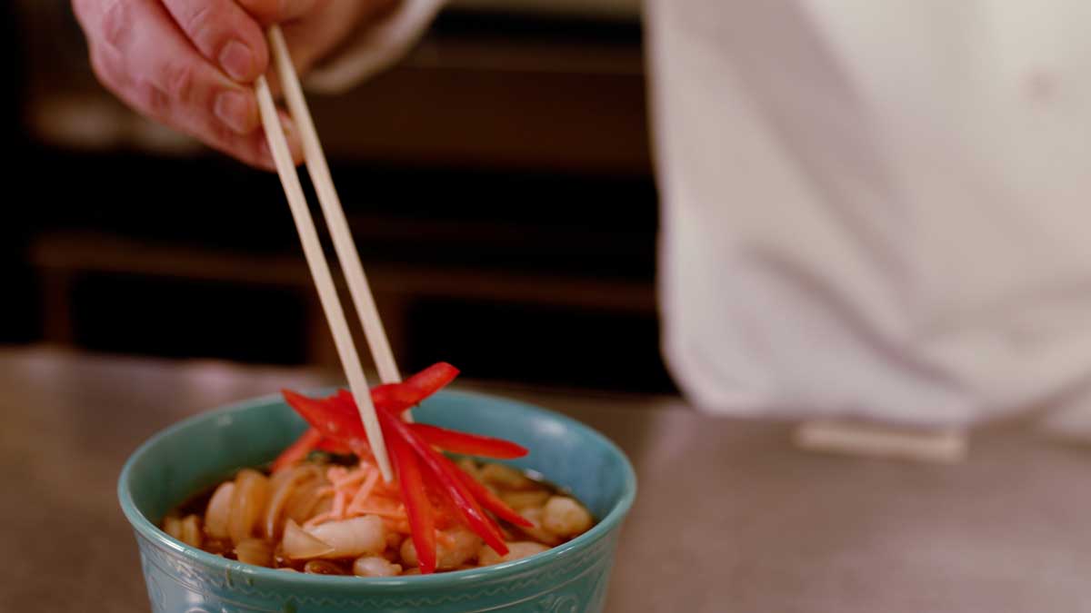 Person adding sliced red peppers to the top of a noodle bowl