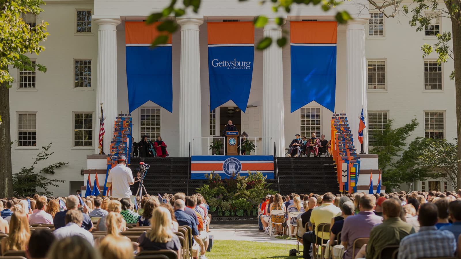 Gettysburg College banners flying from Penn Hall above Beacham Portico during ceremony with colorful audience