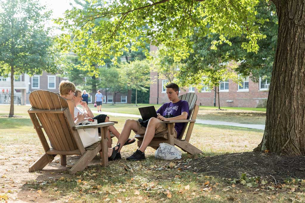 Student sitting in an Adirondack chair on campus using a laptop computer
