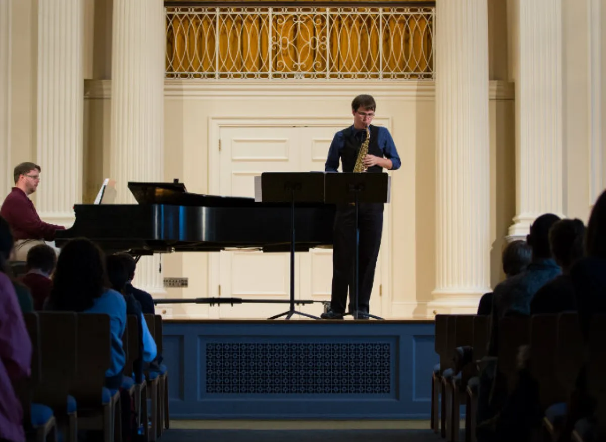 A speaker stands at a podium in a recital hall while a pianist sits at a grand