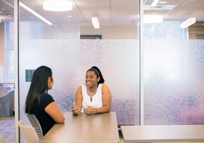Two students talking at a table in a glass-walled room