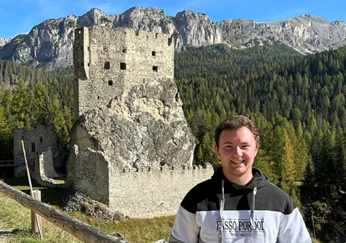 A student poses beside a historic stone ruin with mountains behind.