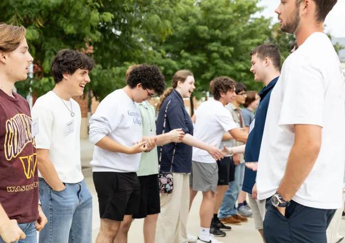 Students stand in two lines outdoors, greeting one another on a tree-lined campus path.