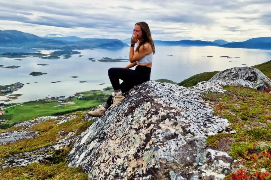 female student sits on rock on mountain with village, body of water, and large mountains in backround