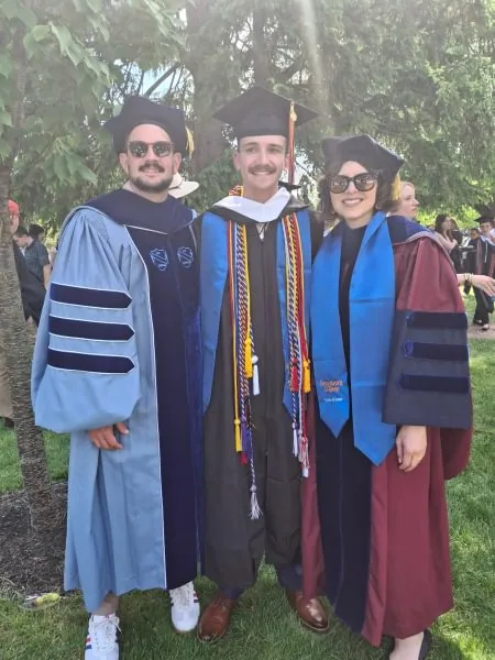 male student in commencement regalia stands between male and female professors