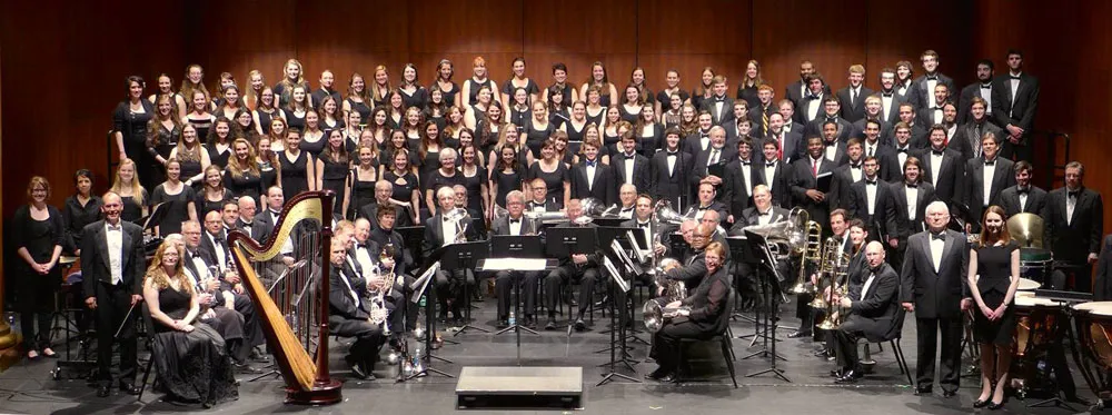 A large choir and orchestra pose together on a concert stage, with a harp, brass, woodwinds, and strings visible in the ensemble.