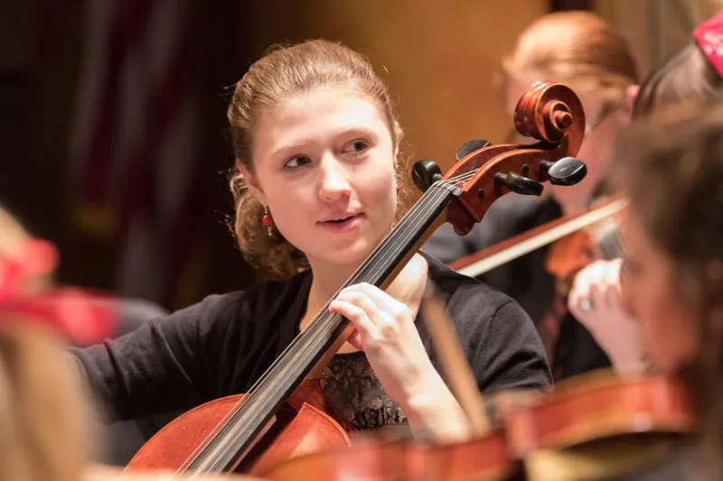 A cellist performs in an orchestra, surrounded by other string players in a warmly lit concert setting.