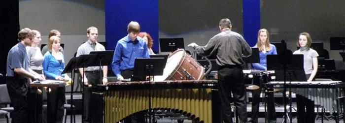 A percussion ensemble rehearses onstage, with musicians gathered around marimbas, drums, cymbals, and music stands while a conductor leads them.