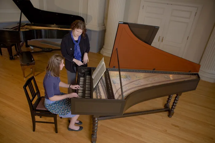 Instructor guides a student playing a harpsichord in a music room.
