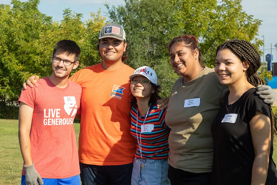 Five students volunteers pose together outdoors at a community service event.  Intellectual wellness: A student presents a project to attendees during an indoor campus showcase.