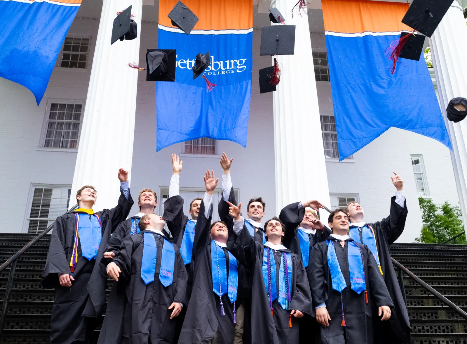 Graduates in black academic gowns and blue stoles celebrate on the steps of the Pennsylvania Hall as graduation caps fly overhead.