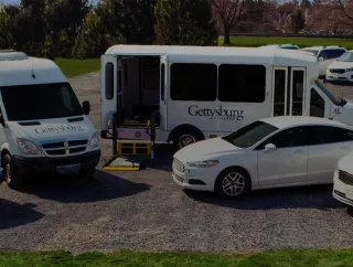 A lineup of Gettysburg college white passenger vans and cars is parked side by side in a lot.