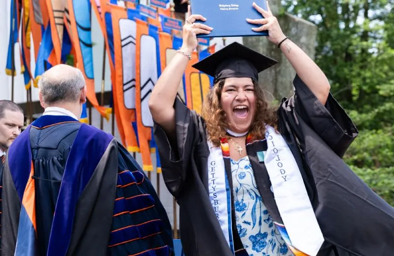 A graduate smiling and holding her diploma above her head.