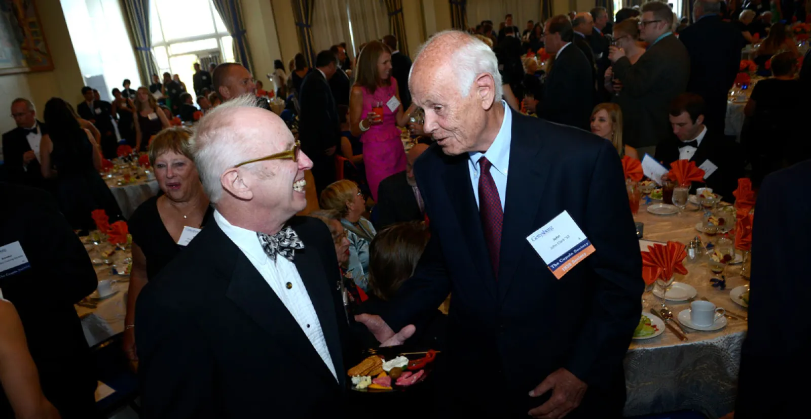 two male alumni speak with busy dinner event in background