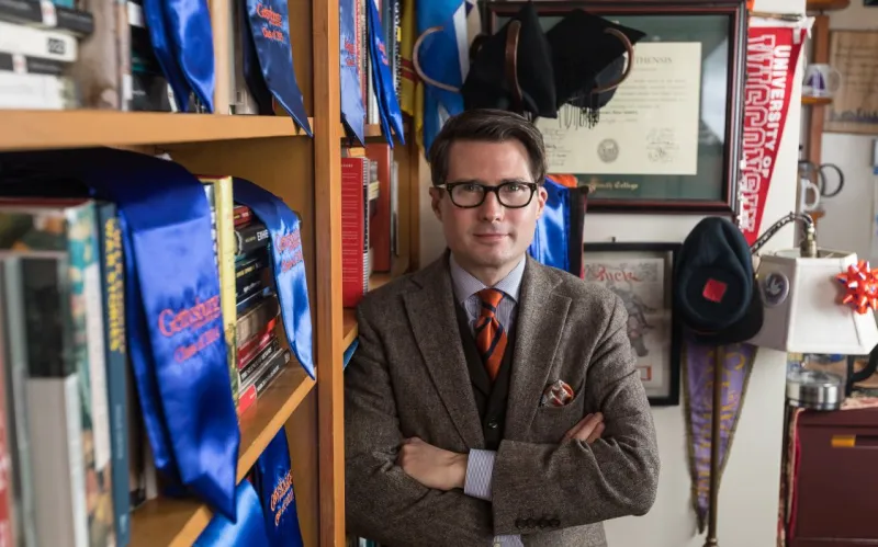 male prof poses leaning on bookcase